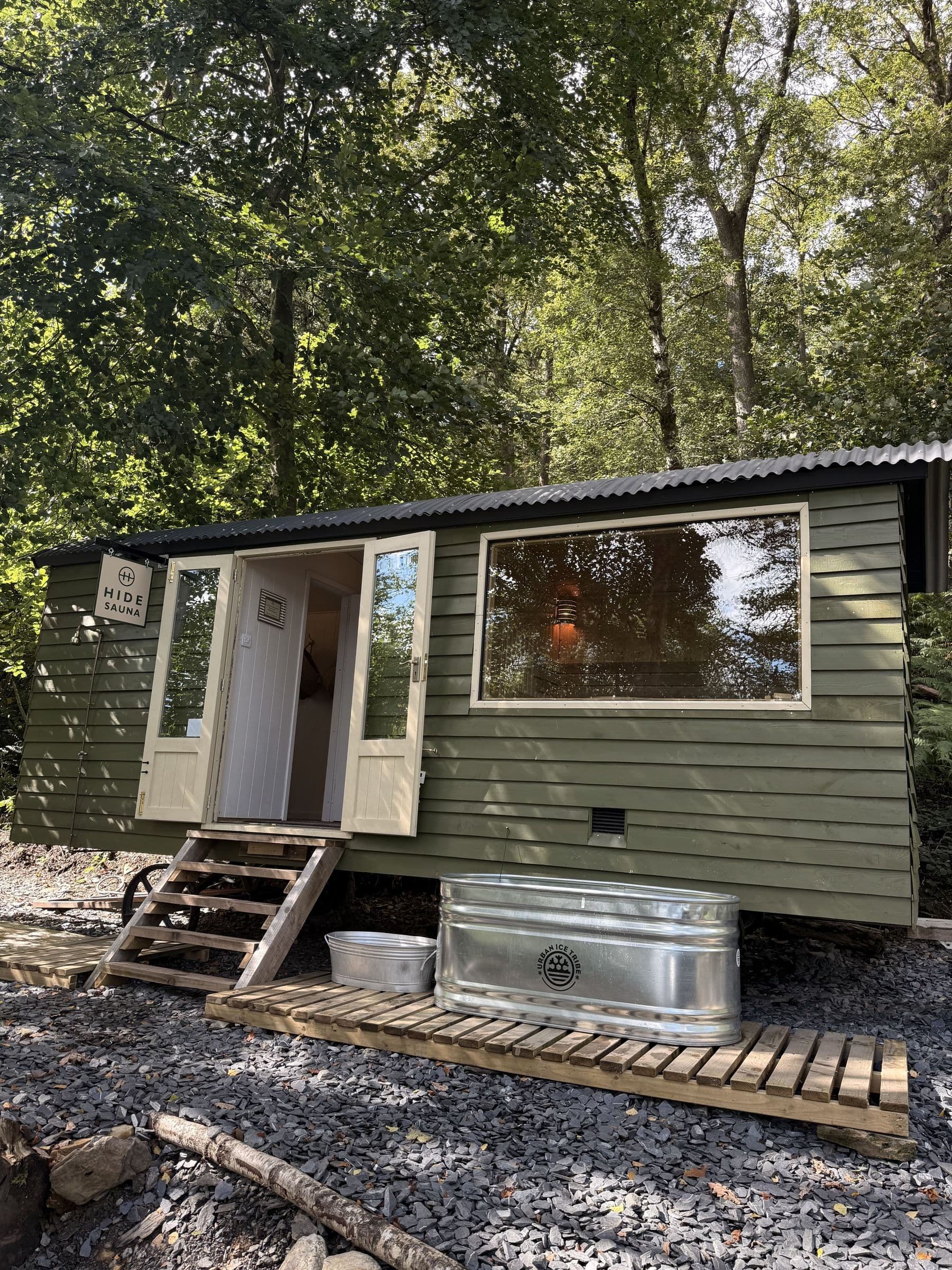 Hide Sauna's green cabin among the trees, its doors open beside the hanging HIDE SAUNA sign