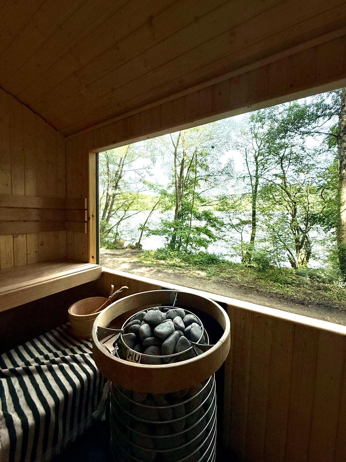 Inside the sauna: the stone heater and a striped towel on the pine bench, the wide window looking out through trees to the lake