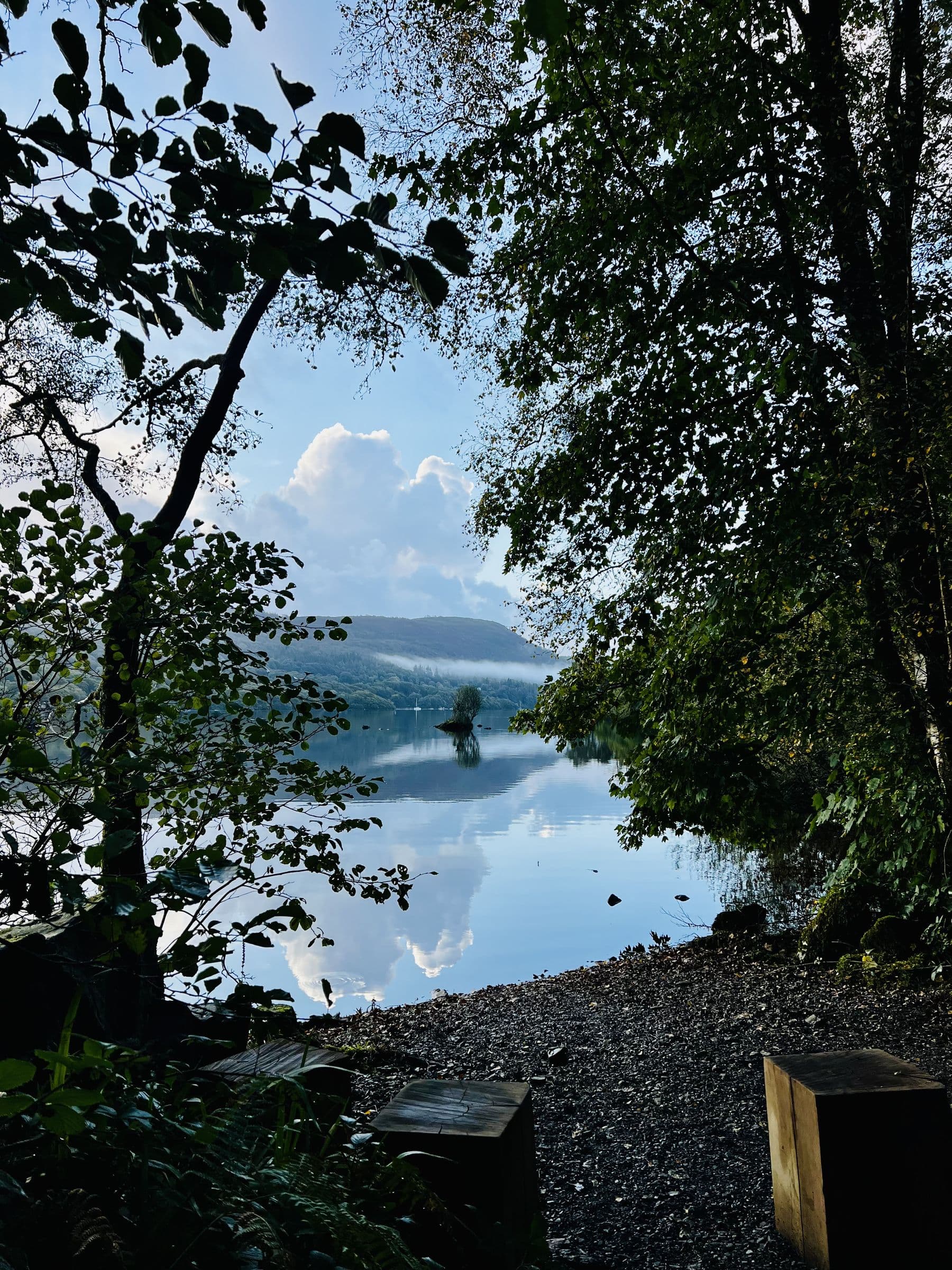 Still Windermere between dark trees, mist on the far hills and clouds mirrored in the water, with stump seats on the stony shore