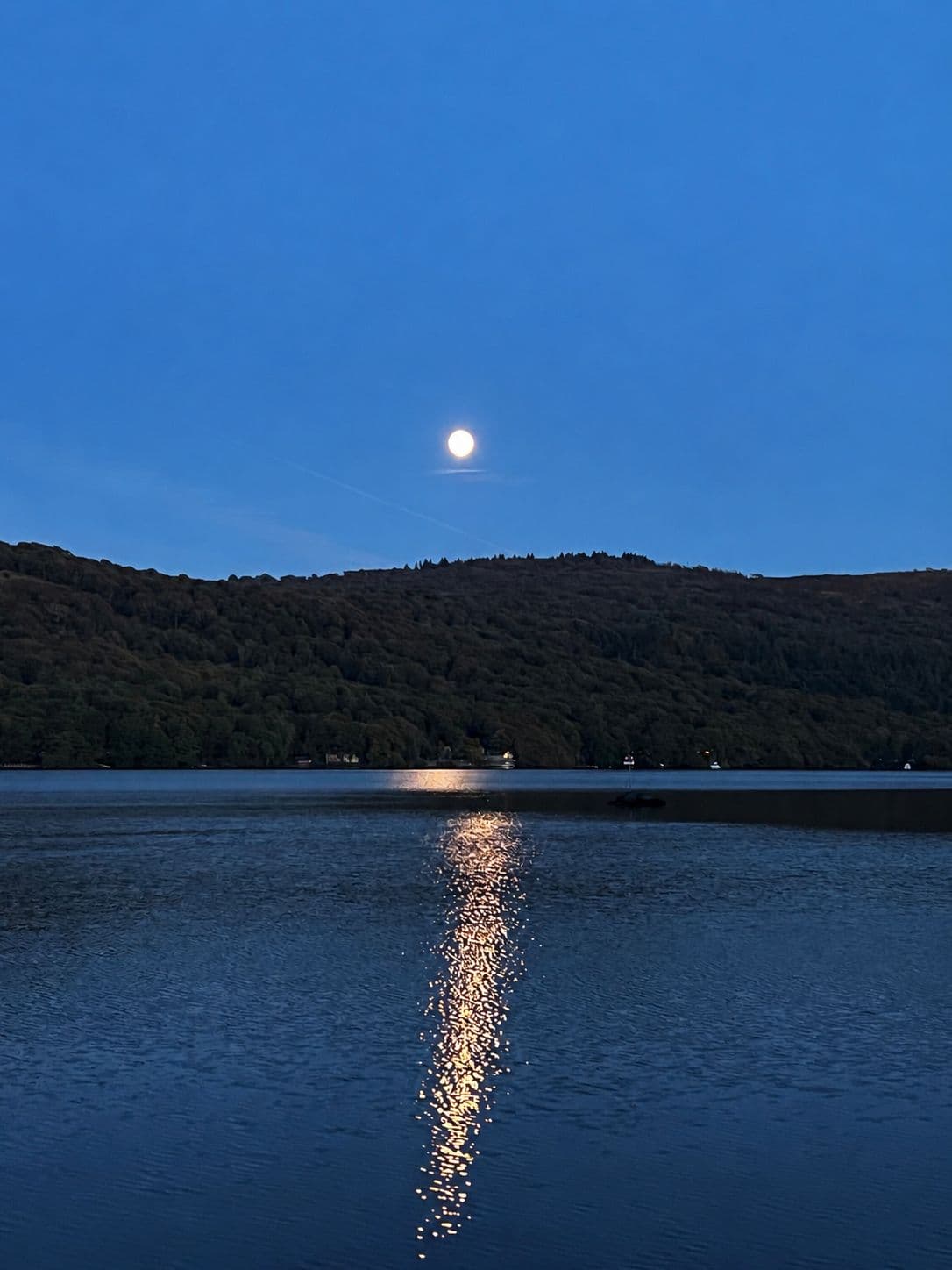 A full moon over the wooded hills at blue hour, its path of light across Windermere