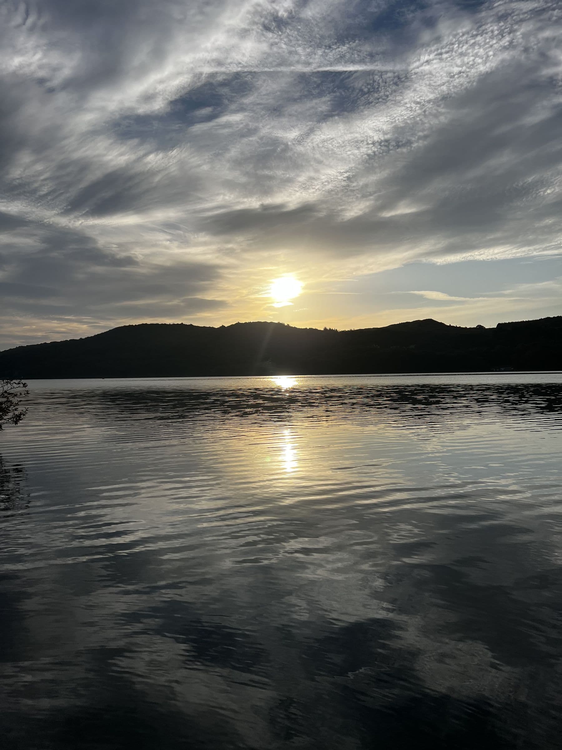 The sun setting behind the fells over Windermere under streaks of cloud, its light reflected on the water