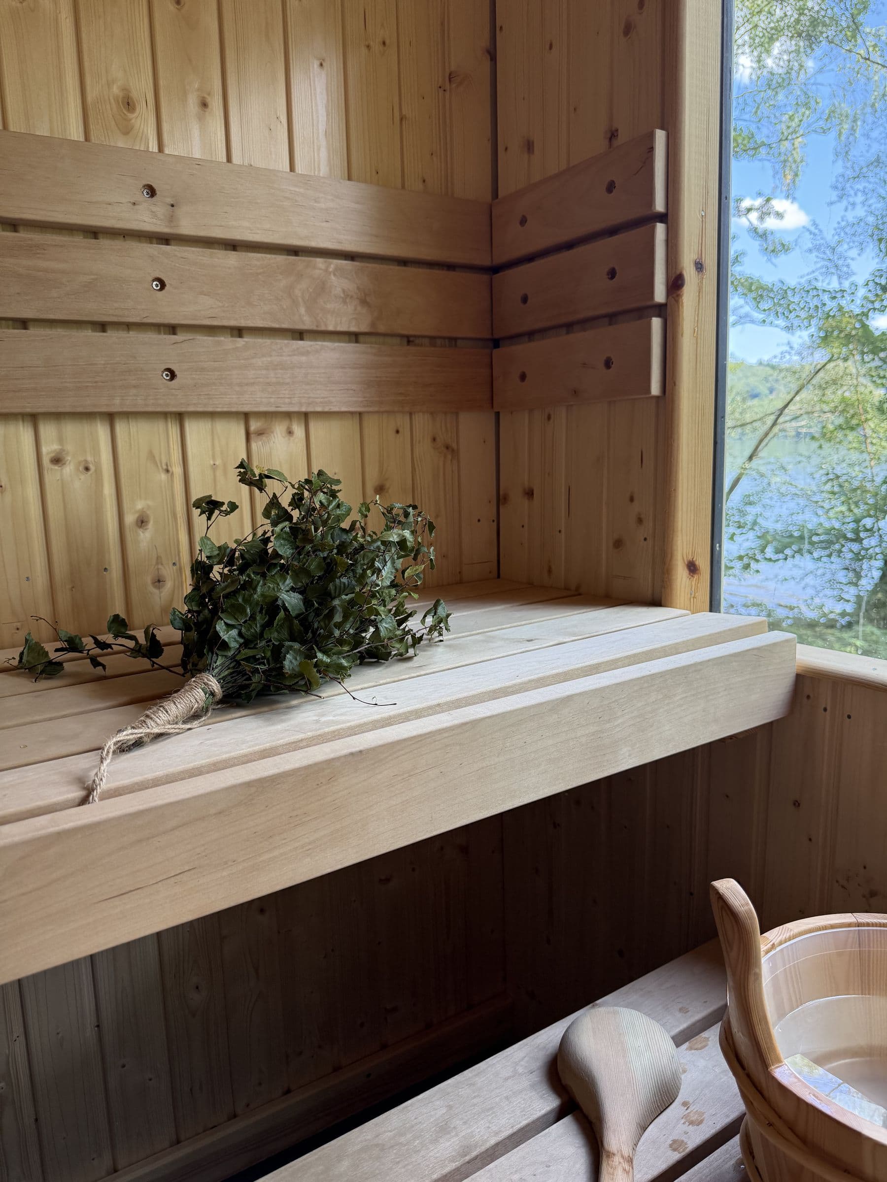 A birch whisk on the pale pine bench inside the sauna, a ladle and wooden bucket below, the lake through the window
